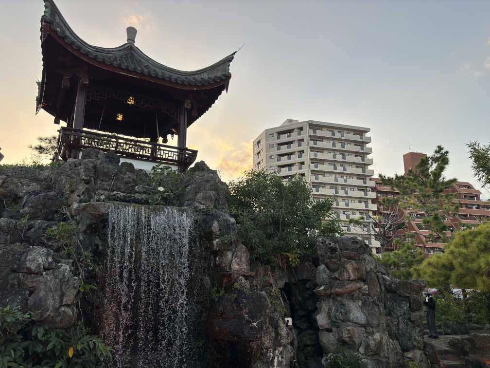 Fukushuen garden, looking at a waterfall with a balcony on top of it. Apartment buildings can be seen in the background